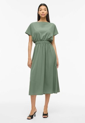 Kobieta w midi sukience w kolorze szałwiowym z krótkimi rękawami i elastyczną talią, stojąca i zwrócona przodem na jednolitym białym tle.