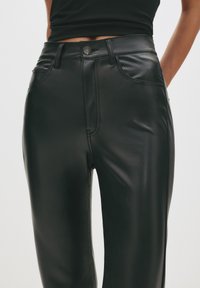 Žena oblečená v čiernych nohaviciach z umelého koženého materiálu s vysokým pásom, ktoré majú bočné a predné vrecká, skombinovaných s čiernym cropped topom.