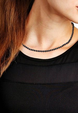 Collier en perles noires avec des perles rondes disposées de manière régulière, porté à la base du cou, contre un haut noir. Texture lisse et design simple.