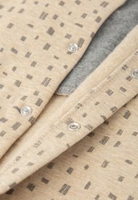 Tejido beige con estampado rectangular gris oscuro, que presenta botones de presión plateados. Textura suave con un forro gris en capas visible debajo.