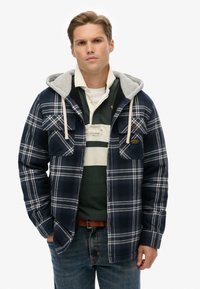 Marineblåternet skjortejakke med grå hætte, to brystlommer, knaplukning og kontrastfarvede hvide linjer. Båret over en grøn polo skorte og jeans.