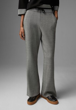 Person iført grå ribstrikkede bukser med vide ben og snor i taljen, sorte sneakers med brune såler og sort langærmet top med grå manchetter.