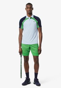 Polo da uomo in azzurro chiaro con dettagli in blu navy e verde, con colletto e maniche corte. Indossato con pantaloni corti verdi e scarpe nere.