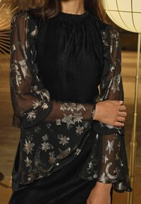 Rochie din catifea neagră cu mâneci transparente negre, împodobite cu modele de stele argintii și lună în formă de semilună; manșete volanate și decolteu plisat.