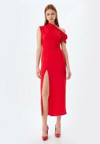 Vestido rojo de un hombro con detalles drapeados y una abertura hasta el muslo. Fabricado en tela suave; combinado con zapatos de tacón alto rojos.