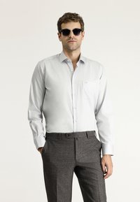 Camicia grigia chiaro a maniche lunghe con bottoni e taschino sul petto, abbinata a pantaloni a quadri scuri. Gli occhiali hanno lenti scure.
