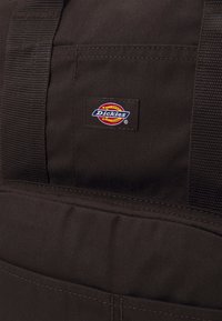 Bolsa de lona marrón con forma rectangular, fabricada con un tejido resistente, costuras reforzadas y un destacado parche con el logo de Dickies.