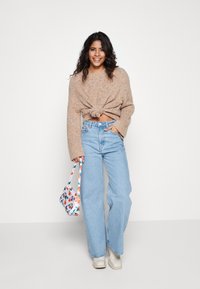 Maglione lavorato a maglia color nocciola con un nodo in vita, abbinato a jeans chiari e morbidi. Tiene in mano una borsa a fantasia floreale e indossa scarpe chiare.