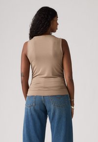 Beige ermeløs topp med en tettsittende design, teksturert stoff og rynkede detaljer bak, sammen med blå vide bukser.
