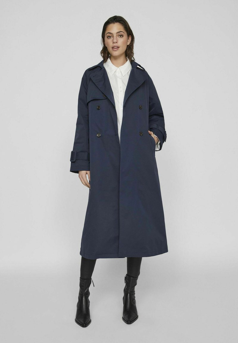 Manteau de trench navy à double boutonnage, avec de grands revers et des manches longues. Présente des accents de boucle sur les manches et des poches latérales.
