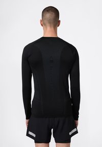 Mann mit kurzen Haaren, der ein schwarzes, langärmeliges Sportshirt mit strukturiertem Rückenpanel und schwarze Shorts mit reflektierenden Streifen trägt, steht mit dem Rücken zur Kamera.