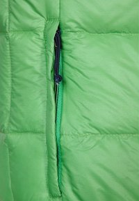 Tkanina jasně zelené nadýchané bundy s viditelným zipem na kapse, modrým jezdcem zipu a kontrastní vnitřní látkou; hladká, prošívaná textura.