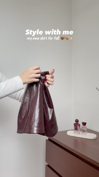 Hände mit roten Nägeln halten einen glänzenden burgunderfarbenen Lederrock neben einer Holzkommode mit zwei Parfümflaschen auf einem weißen Tablett.