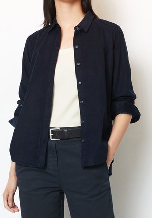 Mujer con una camisa azul oscuro abotonada sobre una camiseta blanca, pantalones negros y cinturón negro, una mano en el bolsillo.