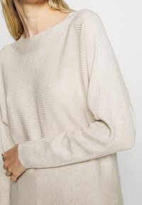 Helles beige, geripptes Strickpullover mit weitem, rundem Halsausschnitt und lockeren langen Ärmeln. Glatte Textur, leicht lockere Passform, minimale Details.