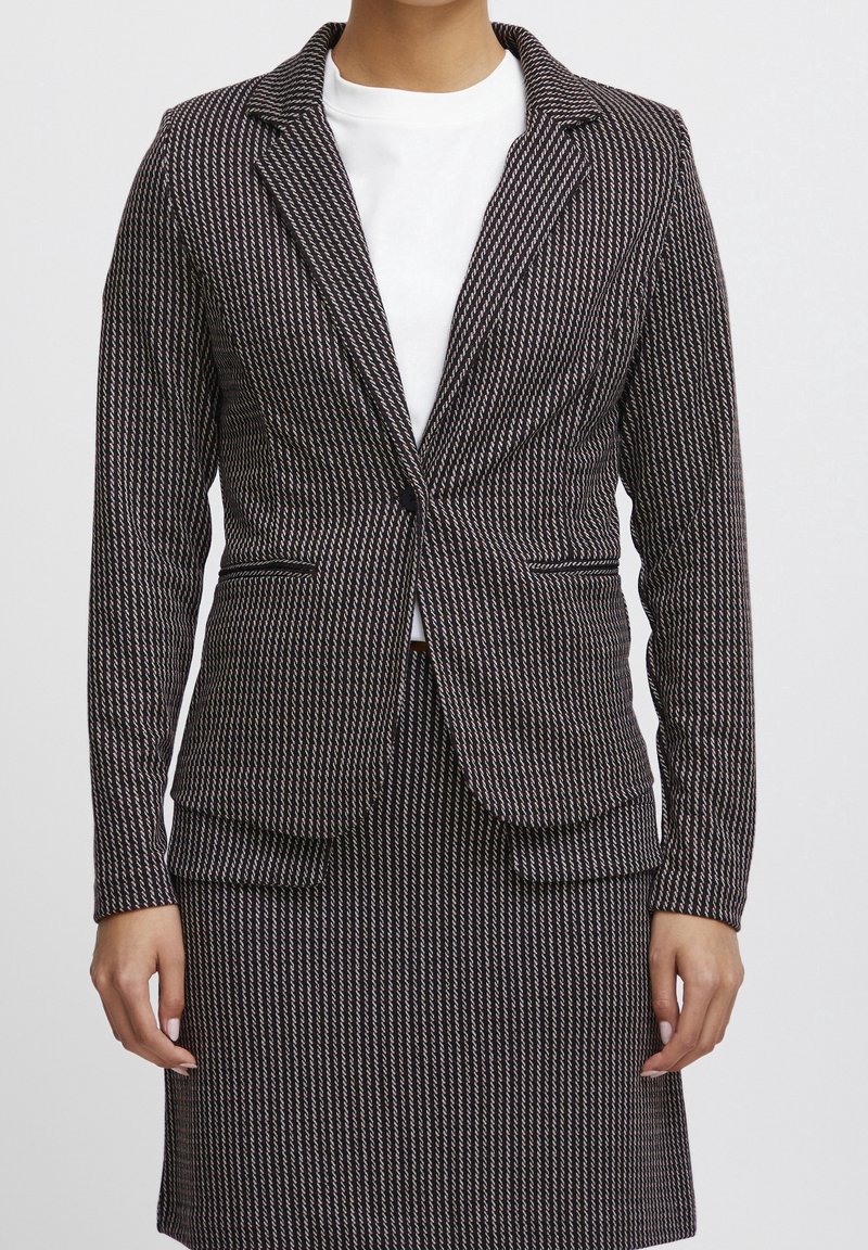 Femme portant un blazer ajusté à motif noir et blanc et une jupe assortie sur un haut blanc, debout avec les bras détendus le long du corps.