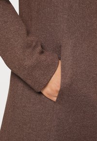 Hnědé šatové šaty vyrobené z texturované pleteniny, s boční kapsou a přiléhavým rukávem, zdůrazňujícími čistý a jednoduchý design.