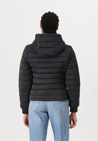 Czarna kurtka puchowa z kapturem, z pikowanymi panelami i prążkowanymi mankietami. Noseny z jasnoniebieskimi dżinsami, pokazany od tyłu.