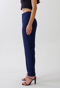 Marinblå skräddarsydda byxor med rak passform och framfåll, tillverkade av ett slätt tyg; bärs med en kort svart topp och remsandaler.