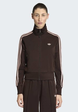 Mujer vistiendo un chándal Adidas marrón oscuro con rayas y logotipos rosas, de pie y mirando al frente contra un fondo liso.