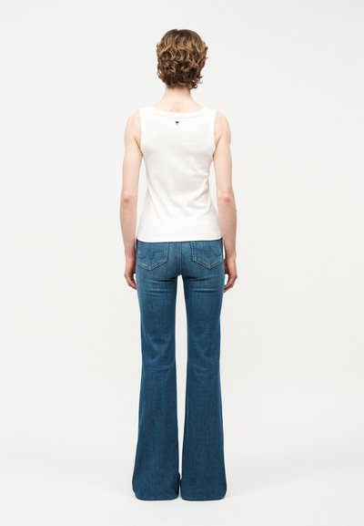 Personne avec des cheveux courts et bouclés portant un haut blanc sans manches et un jean évasé bleu taille haute, debout devant un fond uni.