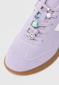 Zamszowy sneakers w kolorze bzu z białymi akcentami, ozdobiony dekoracyjnymi zawieszkami na sznurówki w różnych kolorach, w tym uśmiechniętą buźką i koralikami w paski.