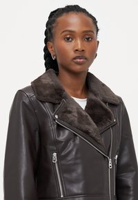Brun læderjakke med en faux fur-krave, asymmetrisk lynlås lukning og sølv hardware detaljer. Glat tekstur med en tætsiddende pasform.