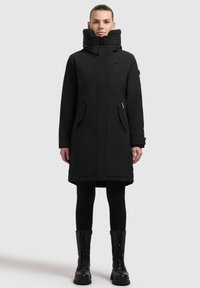 Sort parka med høj krave, der har en struktureret foring, to forlommer og en lige pasform, parret med sorte knæhøje snørebodys.