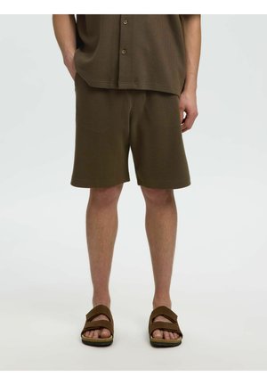 Mężczyzna w oliwkowo-zielonych, teksturze szortach, dopasowanej koszuli i brązowych sandałach, stojący na jednolitym jasnym tle.