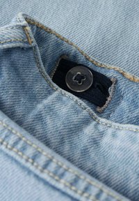 Tessuto di denim azzurro chiaro con un bottone metallico a forma di fiore, cuciture visibili e una toppa di tessuto scuro dietro il bottone.