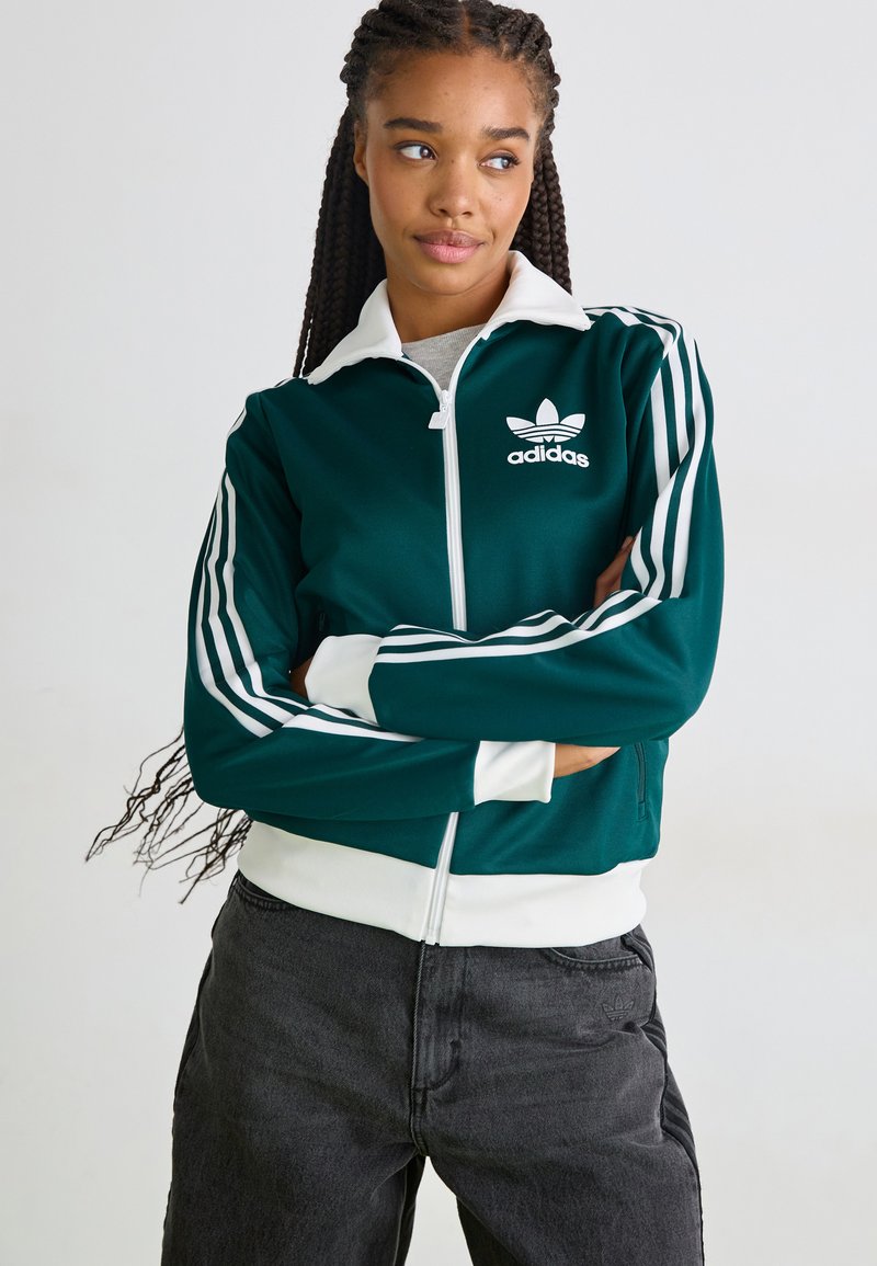 Grüne Zip-Jacke mit weißem Kragen und Bündchen, versehen mit weißen Akzentstreifen an den Ärmeln und einem Adidas-Logo auf der Brust.