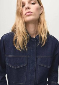 Camicia di denim blu scuro con chiusura frontale a bottoni, colletto appuntito, due tasche sul petto e dettagli di cuciture a contrasto. Texture liscia.