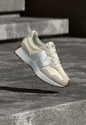 Beige and white New Balance sneaker with silver logo, tan sole, floating above textured gray concrete surface.