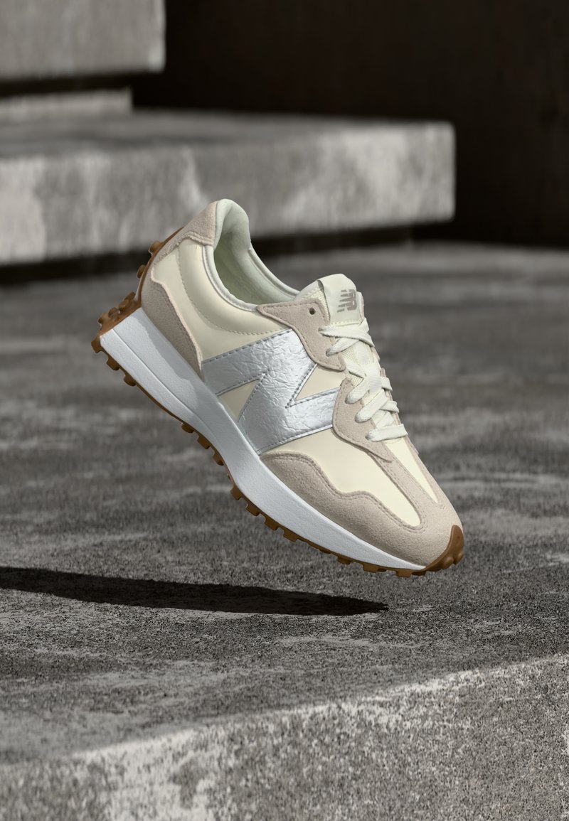 Baskets New Balance beige et blanches avec logo argenté, semelle couleur sable, flottant au-dessus d'une surface en béton gris texturé.