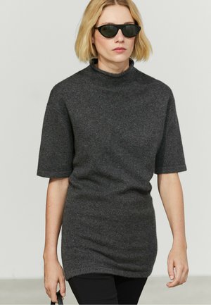 Lyshåret kvinde iført sorte solbriller og en mørkegrå kortærmet højhalset sweater, der holder en sort taske i venstre hånd.
