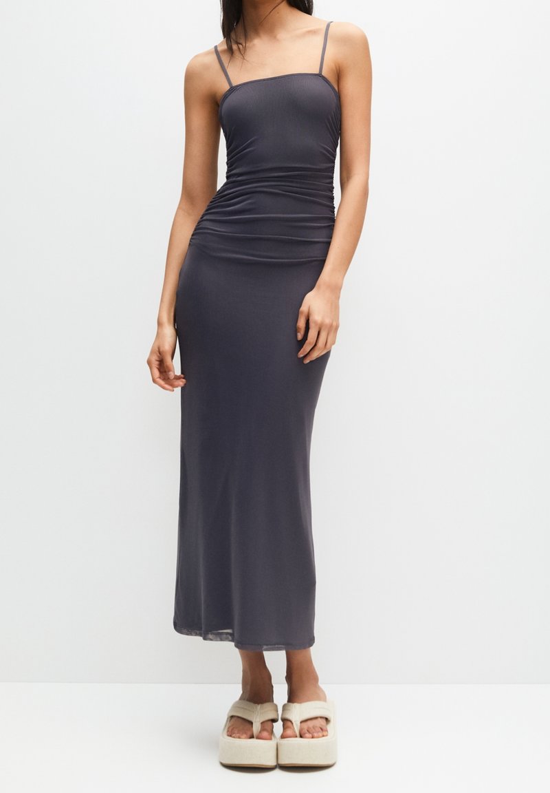 Femme portant une robe maxi ajustée sans manches gris foncé avec fines bretelles et des sandales à plateforme beiges sur un fond blanc uni.