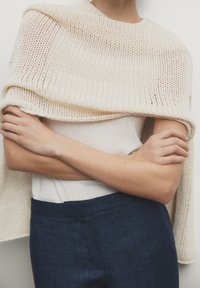 Een gebreide crèmekleurige sjaal hangt over een lichtbeige top, met daaronder zichtbaar donkerblauwe, op maat gemaakte broek. De gestructureerde stof met open patronen is opvallend.