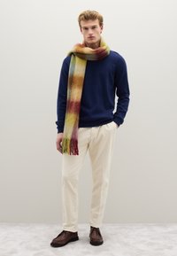 Maglione in lana blu navy, pantaloni in velluto a coste color crema e una sciarpa a frange multicolore in tonalità di rosso, giallo e verde, abbinati a scarpe marroni.