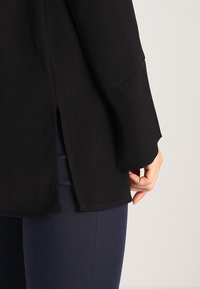 Top negro de manga larga, con una abertura lateral para mayor movimiento. Confeccionado en tejido suave, combinado con pantalones azul oscuro.