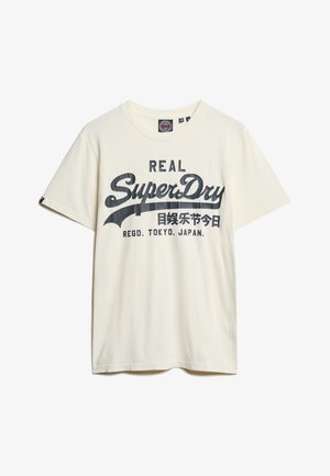 Camiseta de algodón color crema con mangas cortas, que presenta una gran impresión gráfica en azul marino que dice "REAL SuperDry" y texto adicional debajo.