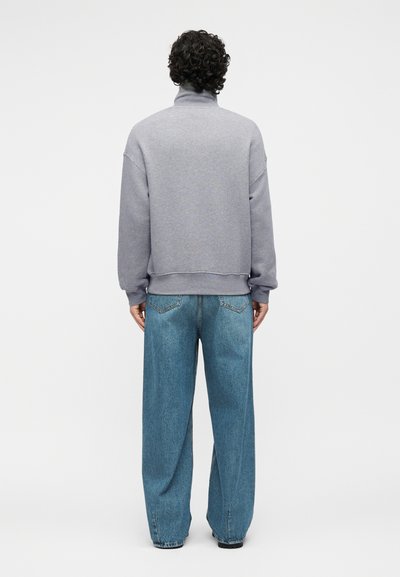 Άτομο με κατσαρά μαλλιά που φοράει γκρι ζιβάγκο φούτερ και φαρδιά μπλε τζιν, στέκεται με την πλάτη προς τα εμπρός σε απλό φόντο.