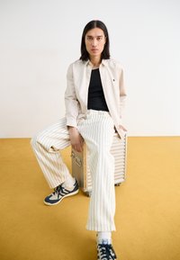 Beige Hemd mit Knöpfen über einem schwarzen Tanktop, kombiniert mit weiß gestreiften Hosen. Blauen und cremefarbenen Sneakers, sitzend auf einem metallischen Koffer.