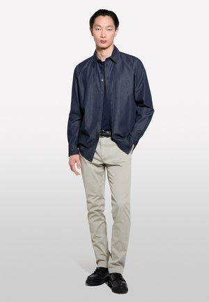 Un jeune homme se tient avec confiance, portant une chemise noire à boutons sur un haut marine, un pantalon beige et des chaussures noires, le tout sur un fond neutre.