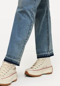 Zapatillas altas beige con parte superior de lona texturizada, que presentan una suela de goma blanca con un borde dentado y detalles en raya roja. Combinadas con jeans de color azul claro.