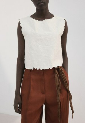 Persona de piel oscura que lleva una blusa sin mangas blanca con textura floral y pantalones de talle alto color óxido, con un accesorio largo de tela marrón.