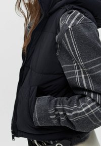 Gilet matelassé avec un corps noir et des manches à carreaux gris. Comprend une capuche, une fermeture éclair à l'avant et des poches latérales. Le tissu texturé met en valeur le design.