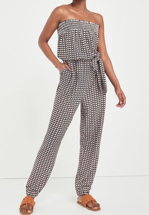 Woman wearing strapless patterned jumpsuit with cinched ankles and a side tie, paired with orange woven slide sandals.
