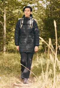 Crna pufasta jakna s kvadratnim uzorkom, crne hlače i sivi planinarski cipele. Ruksak s svijetlim trakama. Prirodna zelena pozadina.