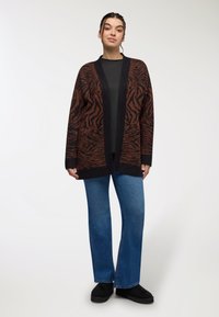 Brązowy i czarny kardigan w zebrę z głębokim dekoltem w serek, długimi rękawami i otwartym przodem; nałożony na czarną bluzkę i zestawiony z niebieskimi dżinsami.