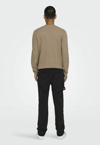 Brun langærmet skjorte med rund hals, parret med sorte cargo-bukser med to baglommer. Modellen vender væk fra kameraet foran en neutral baggrund.